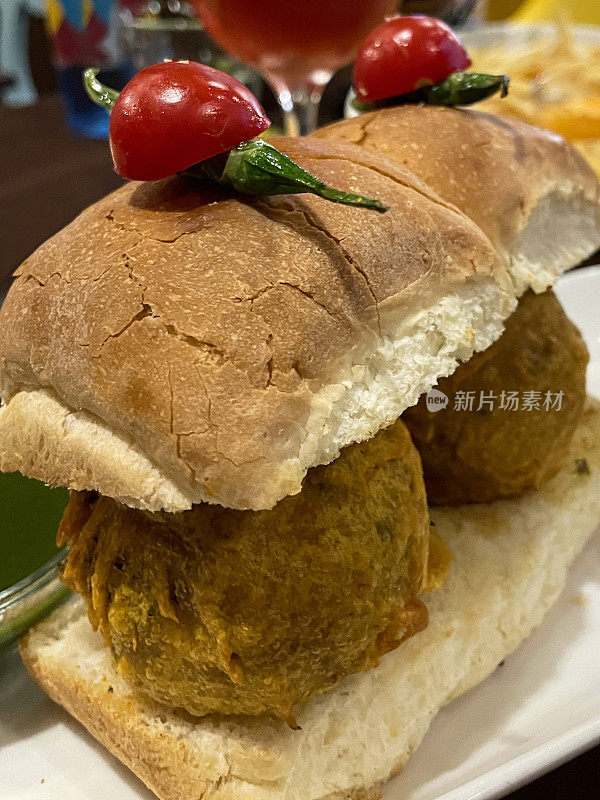 餐厅场景的全帧图像与盘子包含孟买vada pav，油炸土豆饺子在面包包子顶部的绿色辣椒和番茄，重点在前景